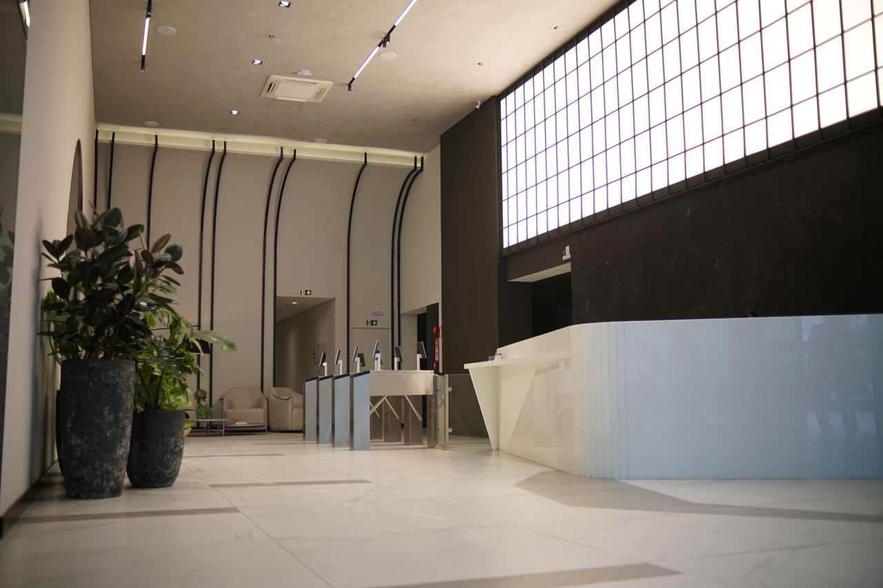 Recepção com catracas de acesso, balcão branco iluminado, parede de blocos de vidro e vasos com plantas